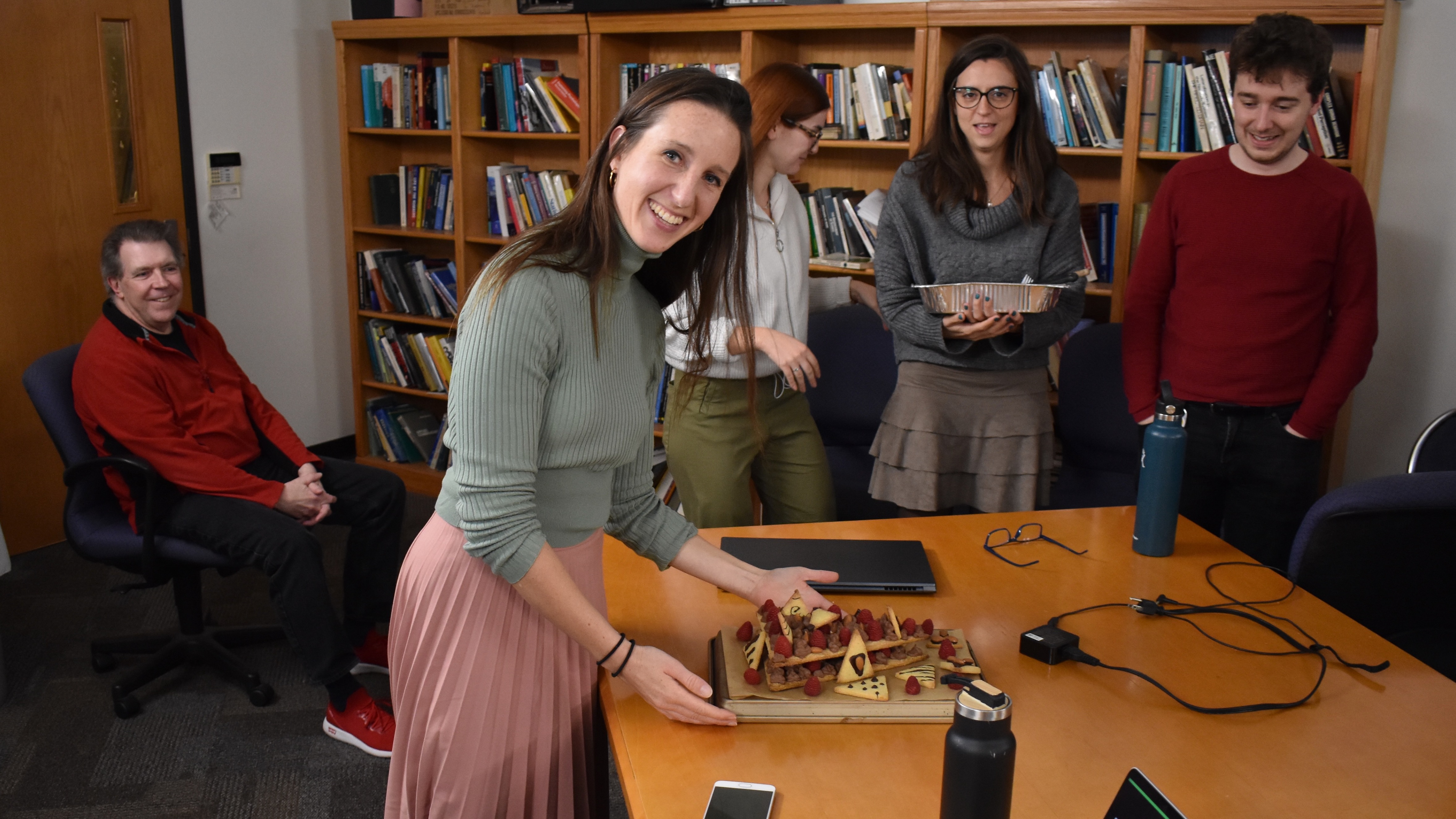 A young woman, happily displaying an array of cookies just gifted to her by her classmates, who stand by in the background.